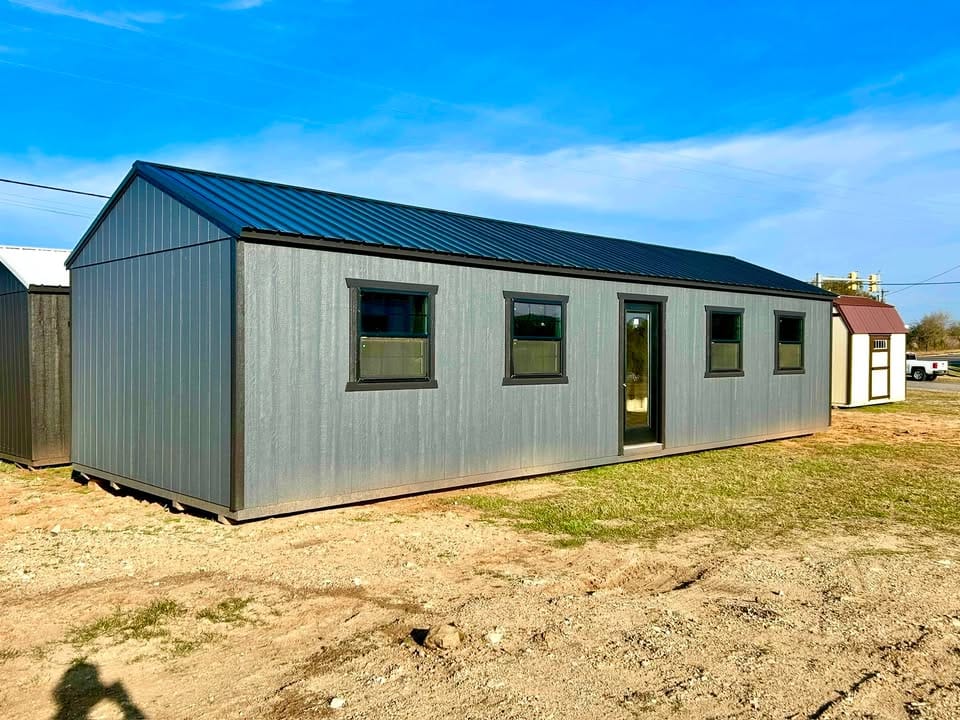 Angled exterior view of a gray 40-foot portable building with wood siding, dark metal roof, four windows, and a centered glass entry door
