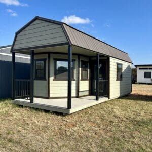 Small cabin-style portable building with front porch, green siding, dark trim, and metal gabled roof displayed outdoors.